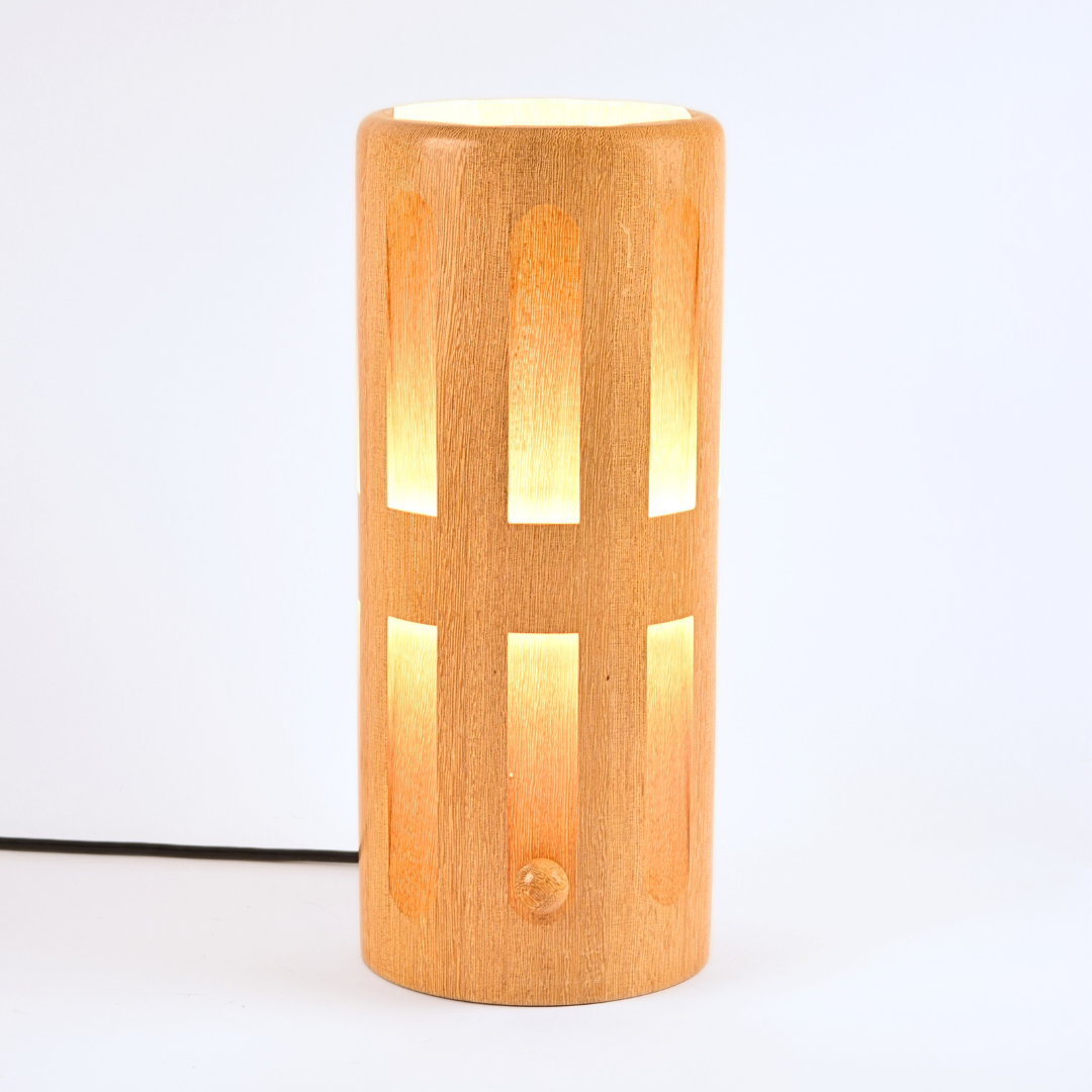 Species: Lati | Dequindre Luminaire
A modern wooden table lamp with a cylindrical body, featuring vertical cutouts that reveal warm yellow light from within. The lamp is made of light natural wood with a visible grain texture and has a small round switch on the front.