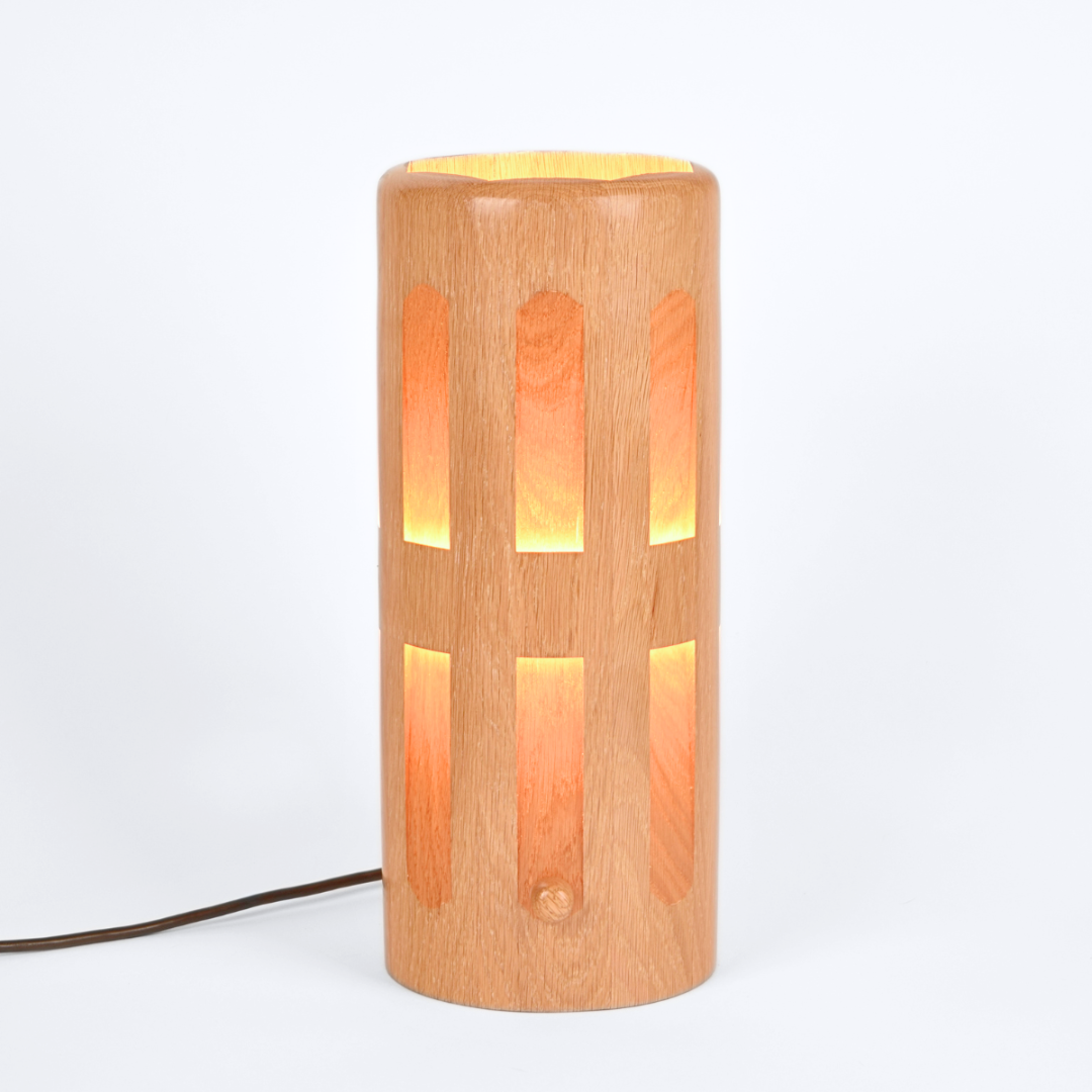 Species: Oak | Dequindre Luminaire
A cylindrical wooden table lamp with a warm light glowing from within, featuring vertical arched cutouts on a light wood-grain finish, shown on a white background with a power cord extending to the left.