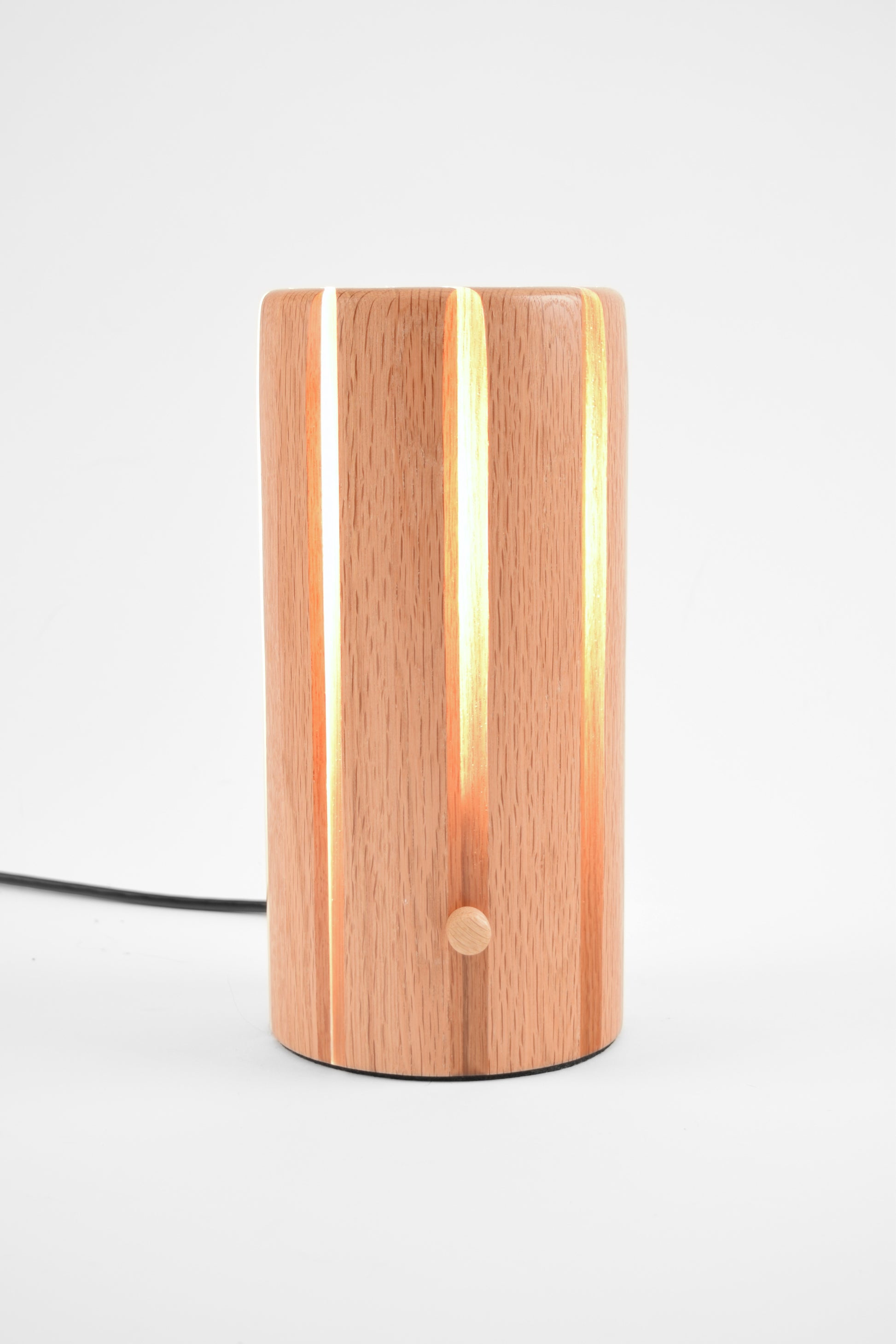 A modern cylindrical table lamp with a light oak wood grain finish, featuring vertical lighted panels that glow with a warm yellow light and a small round button on the front, set against a plain white background.