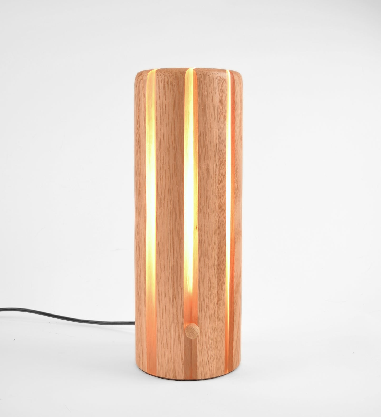 A modern cylindrical table lamp made of vertical wood slats, glowing with warm light from within. A black power cord extends from the base.
