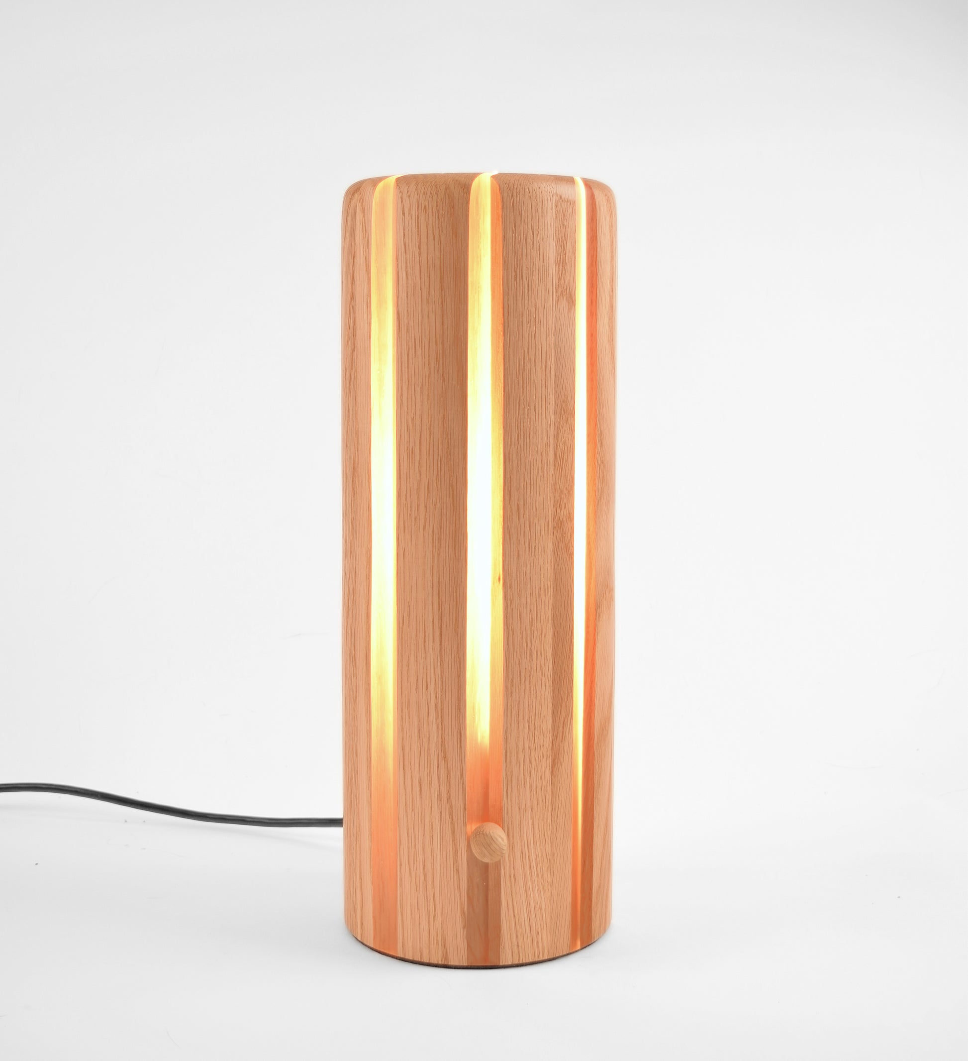 A modern cylindrical table lamp made of vertical wood slats, glowing with warm light from within. A black power cord extends from the base.