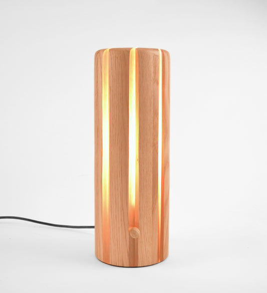 A modern cylindrical table lamp made of vertical wood slats, glowing with warm light from within. A black power cord extends from the base.